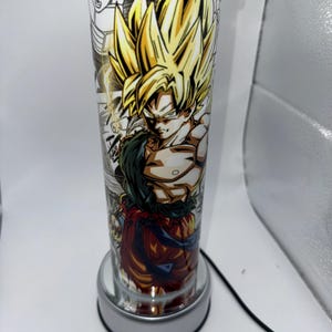 May include: A clear glass tumbler with a colorful anime character design featuring a yellow-haired character with a red and blue outfit. The tumbler is on a silver base with a cord.
