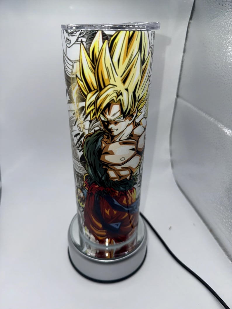 May include: A clear glass tumbler with a colorful anime character design featuring a yellow-haired character with a red and blue outfit. The tumbler is on a silver base with a cord.