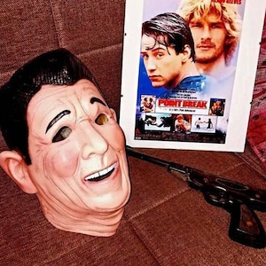 May include: A close-up of a realistic, flesh-toned mask of a man with a wide smile. The mask has black hair and is missing the eyes. The mask is sitting on a brown surface next to a movie poster for "Point Break" and a toy gun.