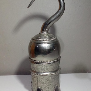 Puede incluir: Un gancho plateado con una hoja curva y una base ornamentada. El gancho es de metal y tiene un diseño decorativo en la base. El gancho es probablemente un accesorio o un artículo decorativo.
