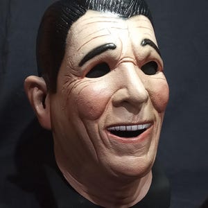 May include: A realistic-looking, smiling face mask made of latex. The mask is painted in a flesh tone and has a black hairpiece. The mask is on a black stand.