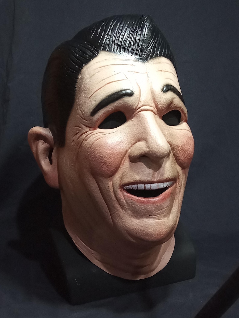 May include: A realistic-looking, smiling face mask made of latex. The mask is painted in a flesh tone and has a black hairpiece. The mask is on a black stand.