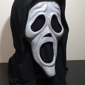 Puede incluir: Una máscara de Scream con una cara blanquecina desgastada y grandes aberturas negras para los ojos. La máscara está colocada contra una capa con capucha negra. La boca de la máscara está abierta en un grito silencioso, un disfraz clásico de Halloween.