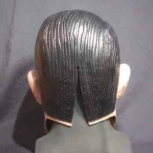 May include: Back view of a black latex mask with a sculpted hairstyle. The mask has a slit in the back for the wearer's head.
