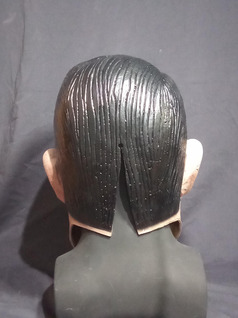 May include: Back view of a black latex mask with a sculpted hairstyle. The mask has a slit in the back for the wearer's head.