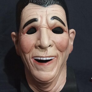 May include: A realistic-looking mask of a man with a wide smile, black hair, and black eyes. The mask is made of a light-colored material and has a textured surface.
