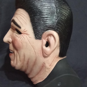 May include: A realistic-looking plastic mask of a man with short, dark hair and a serious expression. The mask is painted in shades of brown and black, and has a detailed sculpt of wrinkles and facial features.