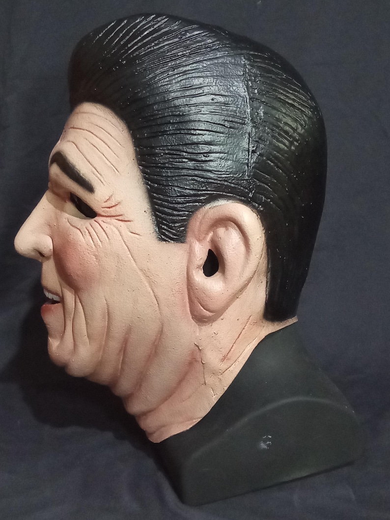 May include: A realistic-looking plastic mask of a man with short, dark hair and a serious expression. The mask is painted in shades of brown and black, and has a detailed sculpt of wrinkles and facial features.
