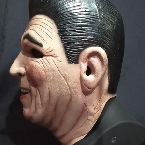 May include: A side profile of a realistic-looking plastic mask of a man with black hair and a light brown complexion. The mask is on a black stand.