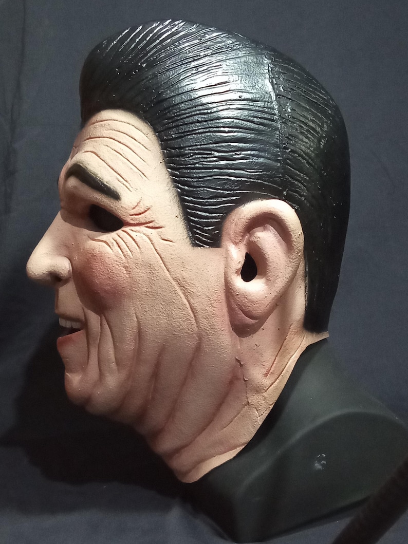 May include: A side profile of a realistic-looking plastic mask of a man with black hair and a light brown complexion. The mask is on a black stand.