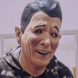 May include: A person wearing a black and white patterned hoodie with a realistic-looking mask of an older man with a smile. The mask has a dark brown complexion and black hair.
