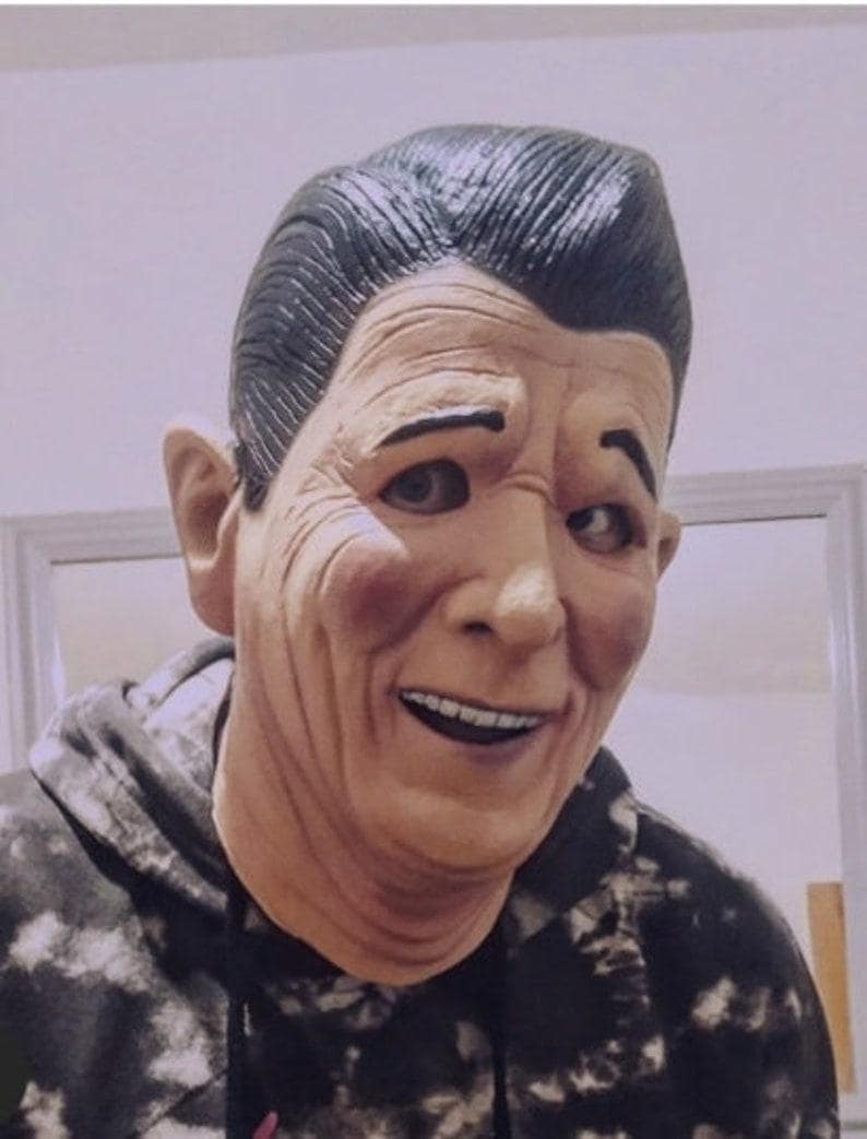 May include: A person wearing a black and white patterned hoodie with a realistic-looking mask of an older man with a smile. The mask has a dark brown complexion and black hair.