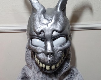 May include: A full-head rabbit mask featuring a metallic silver finish with a textured, weathered surface. The mask has long upright ears, large smooth white eyes, and a wide grin displaying prominent, off-white teeth, attached to a gray faux fur hood.