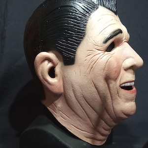 May include: A realistic-looking, detailed, plastic mask of a man's face. The mask is painted in skin tones and has a black wig. The mask is on a black stand.