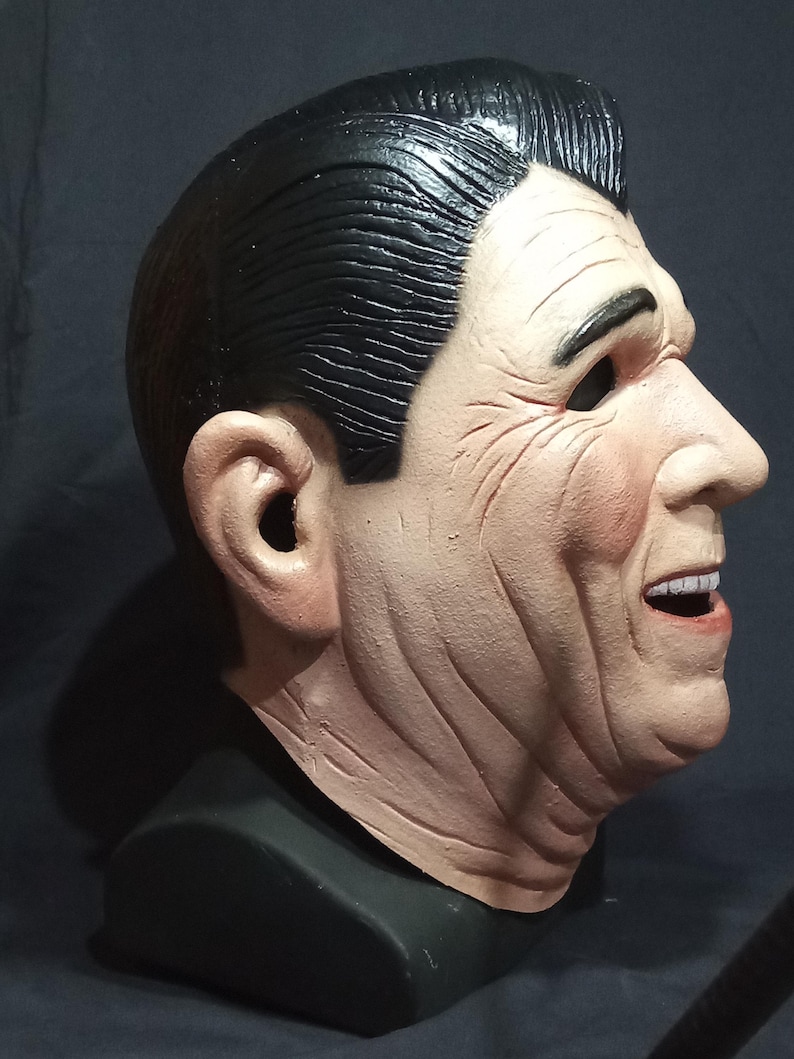 May include: A realistic-looking, detailed, plastic mask of a man's face. The mask is painted in skin tones and has a black wig. The mask is on a black stand.