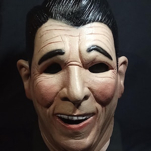 May include: A realistic-looking, white, plastic mask of a man with a black hairpiece. The mask has a wide smile with white teeth and rosy cheeks.