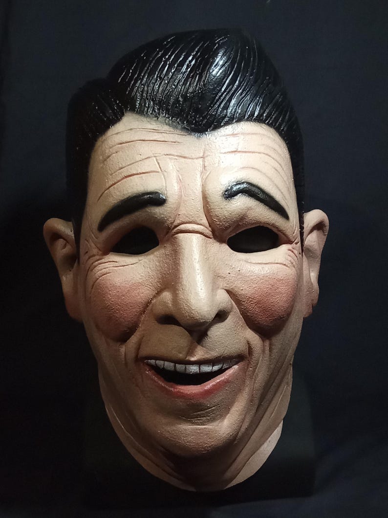 May include: A realistic-looking, white, plastic mask of a man with a black hairpiece. The mask has a wide smile with white teeth and rosy cheeks.