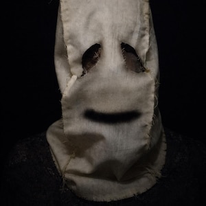 Könnte beinhalten: Eine Maske aus Jute mit Augenlöchern und einem aufgemalten schwarzen Lächeln. Die Maske wird über einem dunkel gefärbten Kleidungsstück getragen, vor schwarzem Hintergrund. Die Ränder der Maske sind grob genäht.