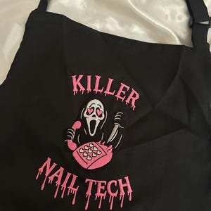 May include: Black apron with adjustable straps featuring pink embroidered text and a graphic. The text reads "KILLER NAIL TECH" with a cartoon ghost holding a phone and knife. The ghost has heart-shaped eyes.