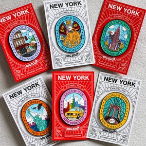 May include: Set of eight enamel pins featuring iconic New York City landmarks. Each pin is on a white card with red and black borders. The pins feature a charging bull, the Empire State Building, the Strand Book Store, Lady Liberty, a yellow cab, and the Brooklyn Bridge. Each card has the text "New York" and "Big Zoo" with the website address.