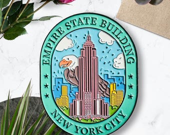 May include: A teal blue enamel pin featuring a cartoon illustration of the Empire State Building, a bald eagle, and the text "Empire State Building New York City".