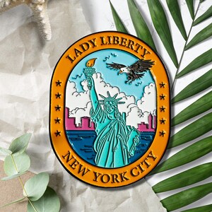 May include: Orange and blue enamel pin featuring Lady Liberty holding a torch, an eagle flying above, and the text "Lady Liberty New York City" in a circle.