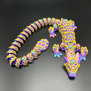 May include: A purple and yellow dragon necklace made of polymer clay. The dragon is segmented and has a long tail. The necklace is made of individual scales that are connected together.