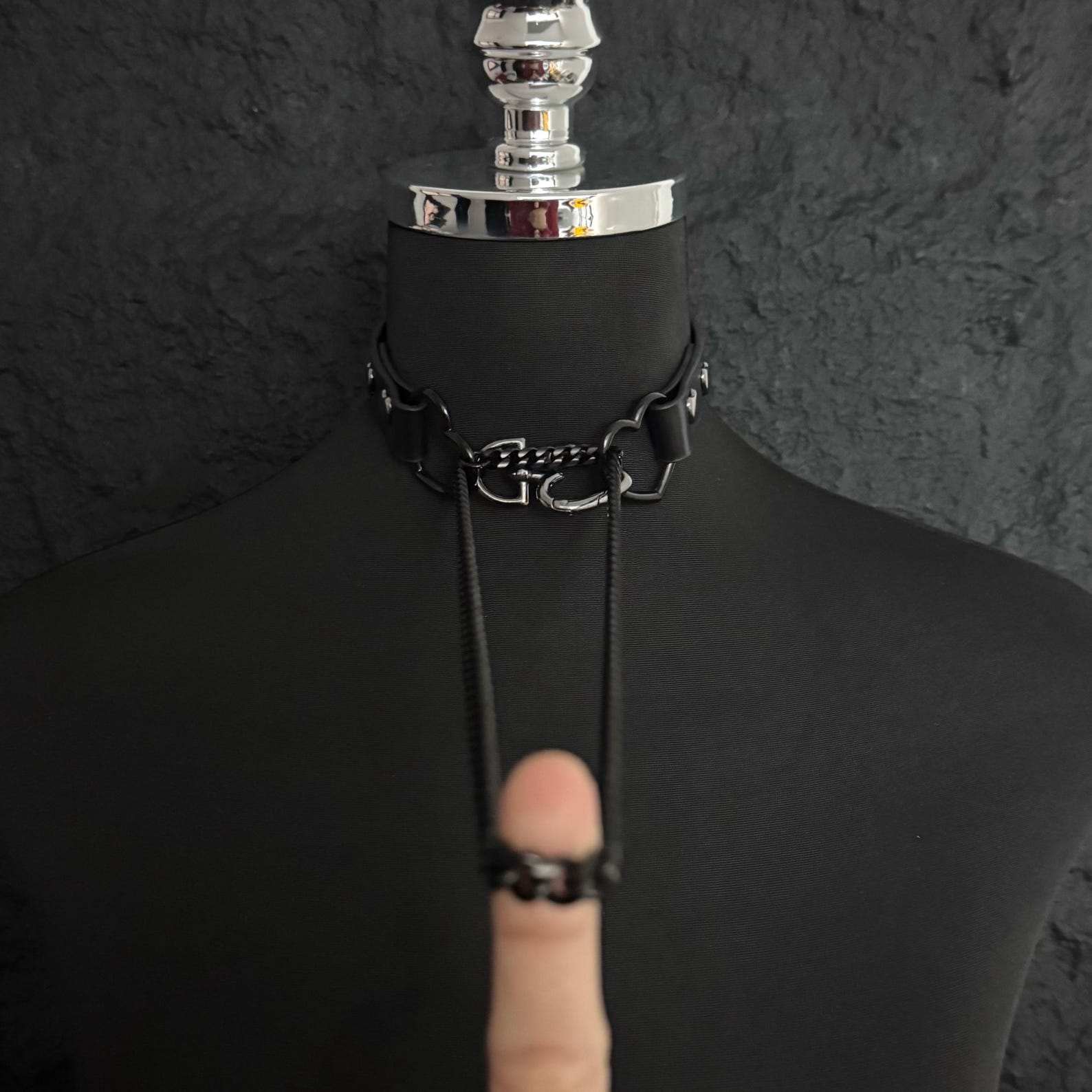 Black leather gothic collar with black chains and heart-shaped rings displayed on mannequin against dark background.