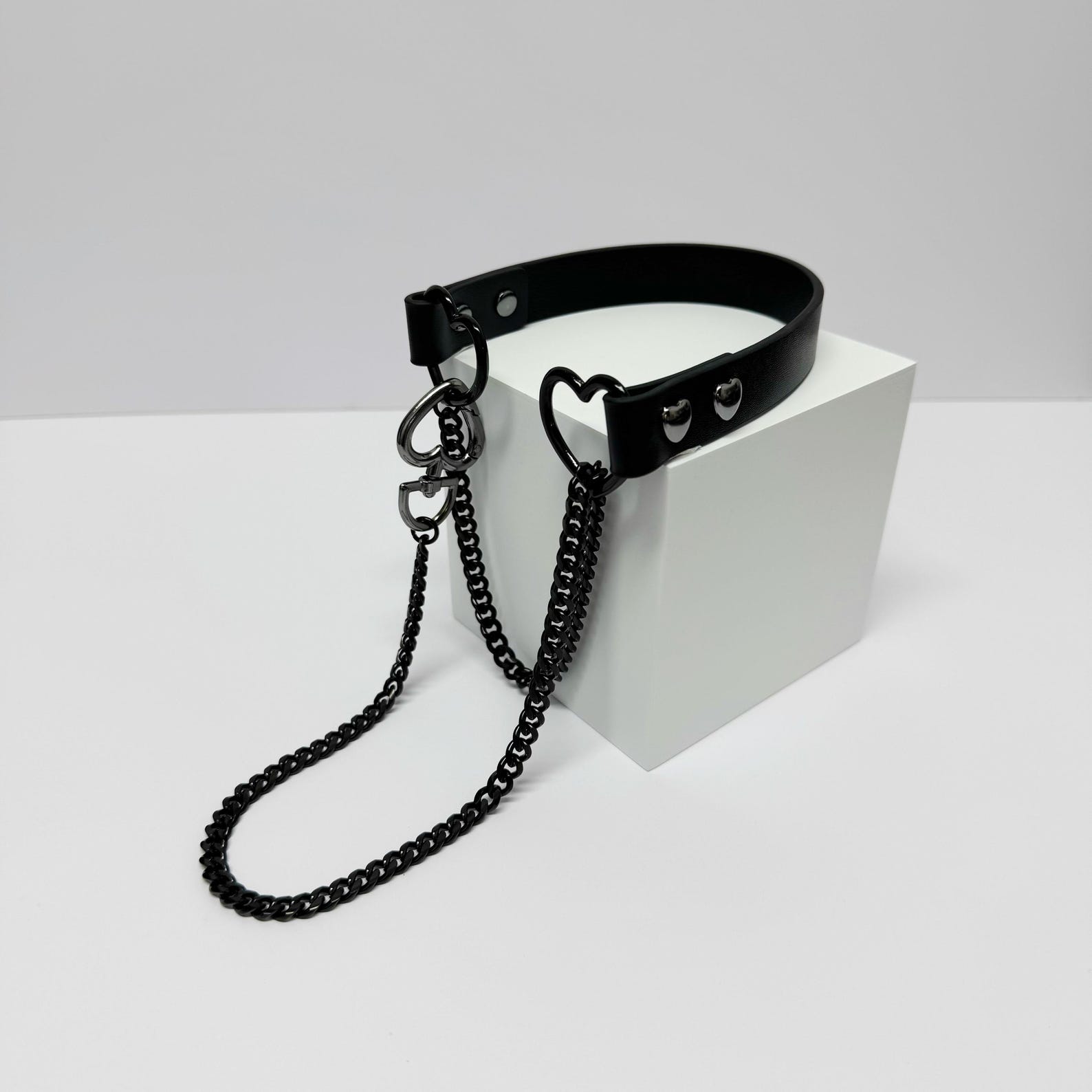 Luxe black leather choker with black chain leash, displayed coiled around white block for dramatic gothic styling.