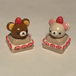 Puede incluir: Dos pequeñas figuritas de arcilla hechas a mano de osos con sombreros de fresa. Los osos están sentados sobre bases cuadradas, marrones, con forma de pastel con glaseado rosa y blanco.
