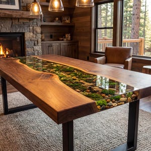 May include: A handcrafted wooden dining table with a river-like design. The center features a clear resin filled with stones, moss, and greenery. The table has a dark metal base and is set in a rustic living room.