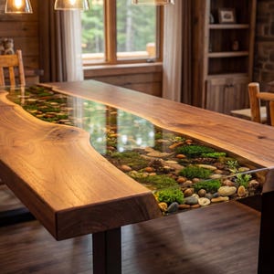 May include: A handcrafted wooden dining table with a clear epoxy resin river running through the center. The river is filled with stones, moss, and small plants. The table has a natural wood grain finish and sits on black metal legs.