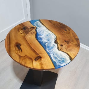 Könnte beinhalten: Runder Holztisch mit einem Fluss aus blauem Kunstharz und weißen Akzenten. Der Tisch hat eine natürliche Holzmaserung und ein schwarzes Metallgestell. Der Tisch hat einen Durchmesser von ca. 76 cm.