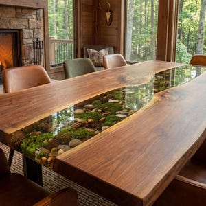 May include: A live edge dining table with a river of clear resin filled with moss, rocks, and greenery. The table is surrounded by leather and upholstered chairs. A fireplace and large windows are in the background.