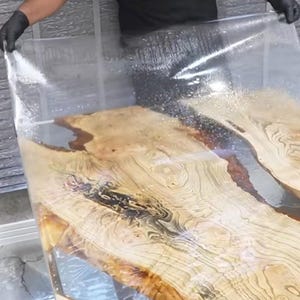 May include: A clear, protective sheet being applied to a wooden table. The table features a light-colored wood grain with dark brown accents and a clear resin finish. The table is rectangular and appears to be handmade.