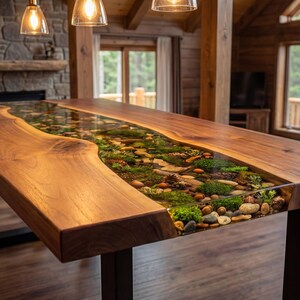May include: A handcrafted wooden table with a river-like design. The table features a clear resin center filled with stones, moss, and greenery. The table is supported by black metal legs and is in a rustic setting.