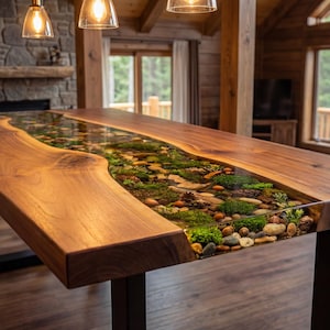 May include: A handcrafted wooden table with a river-like design. The table features a clear resin center filled with stones, moss, and greenery. The table is supported by black metal legs and is in a rustic setting.
