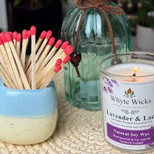 Op de afbeelding: Een transparante glazen kaars in een pot met een wit etiket waarop staat "Whyte Wicks Lavender & Lace Lavender French Essential Oil Promotes relaxation and restful sleep Natural Soy Wax Hand-poured in Co. Antrim Northern Ireland". De kaars is aangestoken en de vlam is zichtbaar. De kaars staat op een geweven oppervlak naast een blauwe en witte keramische luciferhouder gevuld met lucifers.