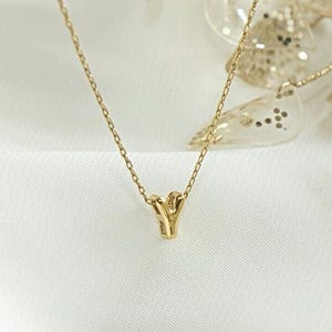 May include: A delicate gold necklace featuring a "Y" pendant. The necklace is set against a white, textured backdrop, with decorative gold leaf accents. The jewellery is elegant and minimalist, suitable for everyday wear or special occasions.