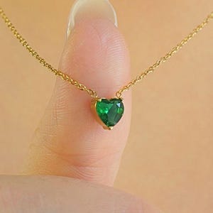May include: A delicate gold necklace featuring a heart-shaped, emerald-green gemstone pendant. The pendant is suspended from a fine gold chain. The necklace is displayed on a person's finger.
