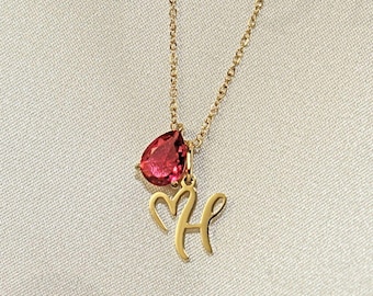 May include: A gold-colored necklace with a teardrop-shaped, red gemstone pendant and a cursive letter "H" charm. The necklace hangs on a delicate chain, set against a soft, white background.