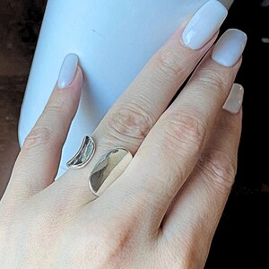 May include: A silver ring featuring a crescent and oval design. The ring is worn on a finger, highlighting its minimalist style. The open design allows for an adjustable fit. The ring's polished surface reflects light, adding to its modern appeal.