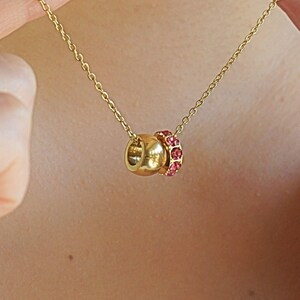 Può includere: Una collana dorata con una catena sottile. Il ciondolo presenta una sfera dorata accanto a un anello di piccole pietre preziose rosa. La collana è tenuta da due mani.