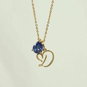May include: A gold-colored necklace with a blue teardrop-shaped gemstone pendant and a heart-shaped letter "D" charm. The chain is delicate, and the pendant hangs in the center, creating an elegant and personalized jewelry piece.