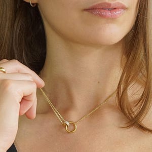 May include: A gold necklace featuring a circular pendant and a fine chain. The image also shows a matching gold ring and small hoop earrings. The jewellery is worn against a dark background.