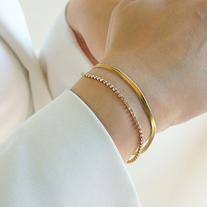 Op de afbeelding: Twee gouden armbanden om een pols. De ene is een dunne, platte band en de andere is een delicate ketting met kleine, heldere stenen. De armbanden worden gedragen om een pols met een witte mouw.