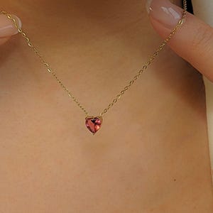 May include: A delicate gold necklace with a heart-shaped, pink gemstone pendant. The pendant hangs from a fine gold chain. The necklace is shown against a skin tone background.