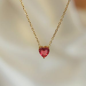May include: A delicate gold necklace featuring a heart-shaped, pink gemstone pendant. The pendant is a vibrant shade of pink, and the chain is a fine, gold link. The necklace is displayed against a soft, neutral background.