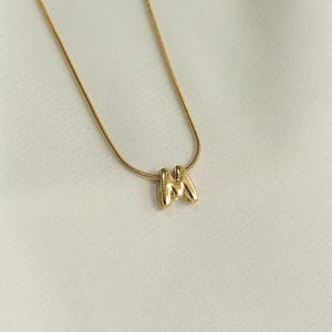 May include: A gold-colored necklace with a pendant in the shape of the letter "M". The pendant is a three-dimensional design. The necklace is displayed on a white, textured surface.