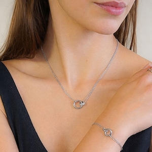Puede incluir: Un conjunto de collar y pulsera plateados. El collar presenta una delicada cadena con un colgante de doble círculo. La pulsera a juego también tiene un diseño de doble círculo. La joyería se usa sobre una blusa negra.